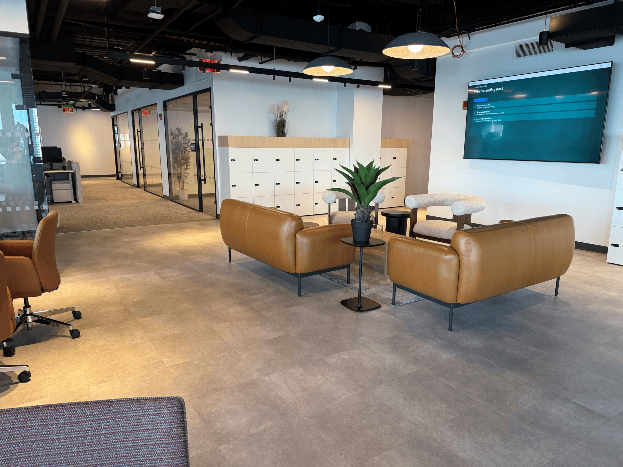Contemporary office collaboration space featuring two tan leather sofas, cushioned lounge chairs, and small black side tables arranged around a central plant. In the background, white storage lockers line the wall beside glass-fronted meeting rooms and office workspaces. A large digital display screen is mounted on the wall, while pendant lighting and an open ceiling create a modern, professional atmosphere.
